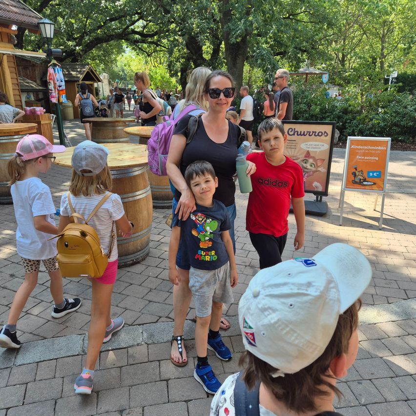 Eine Frau mit Sonnenbrille steht mit drei Kindern vor einem Holzfass und einem Schild. Die Kinder tragen Mützen und Turnschuhe. Dahinter stehen Holzfässer und ein Schild.