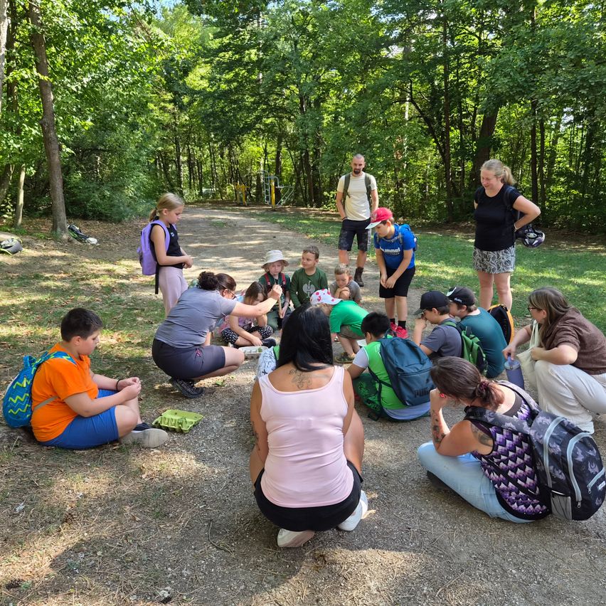 A group of people, including children, are sitting in a circle on the ground in a forest. Some are wearing backpacks.