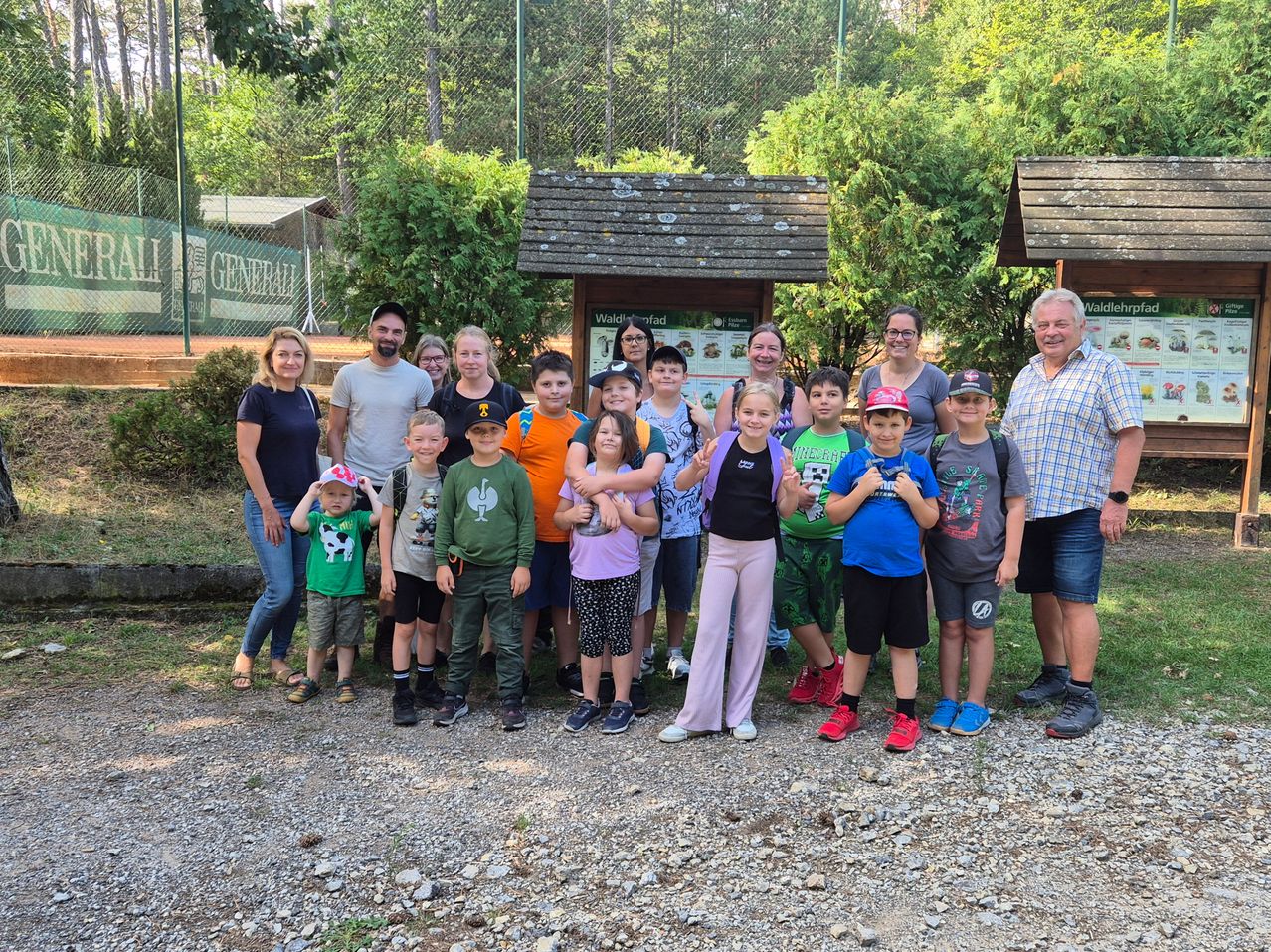 A group of adults and children pose for a photo in a park with a wooden sign and greenery in the background.