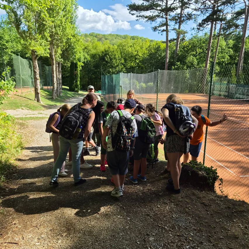 A group of people with backpacks are standing near a tennis court, looking at something on the other side of the fence.