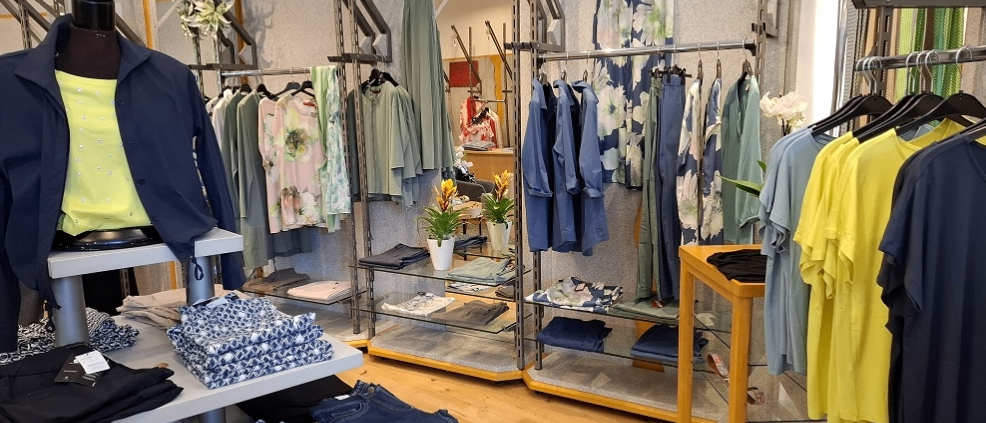 Ein Bekleidungsgeschäft mit verschiedenen Kleidungsstücken, die an Gestellen hängen und auf Regalen gefaltet sind. Blumen- und geometrische Muster sind prominent. Zwei Topfpflanzen stehen in der Mitte.