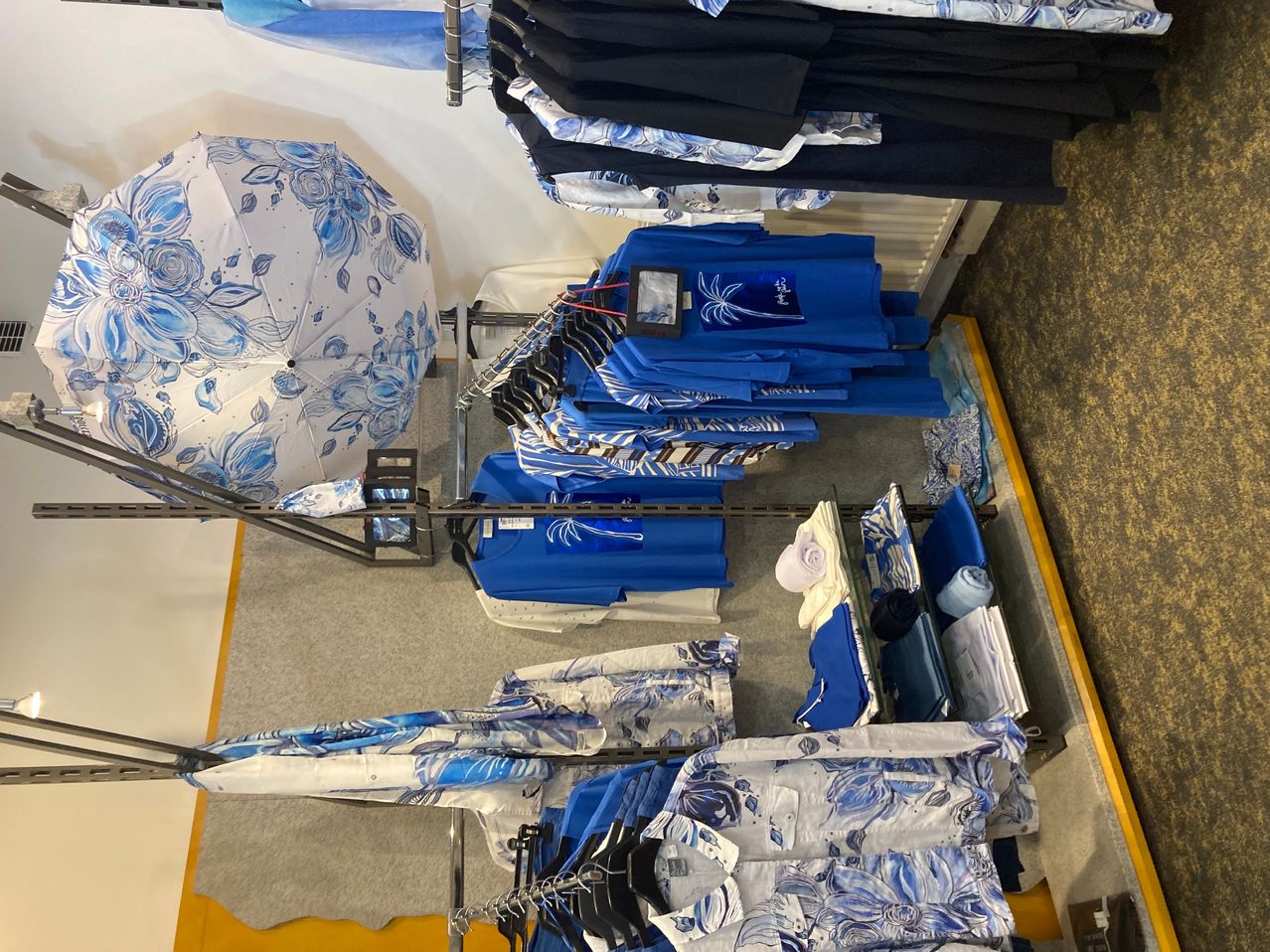 Ein Geschäft zeigt eine Reihe von blauer und weißer Kleidung, einschließlich Hemden, Shorts und einem blauen Blumenschirm. Kleidung hängt an Ständern, und einige sind gefaltet.