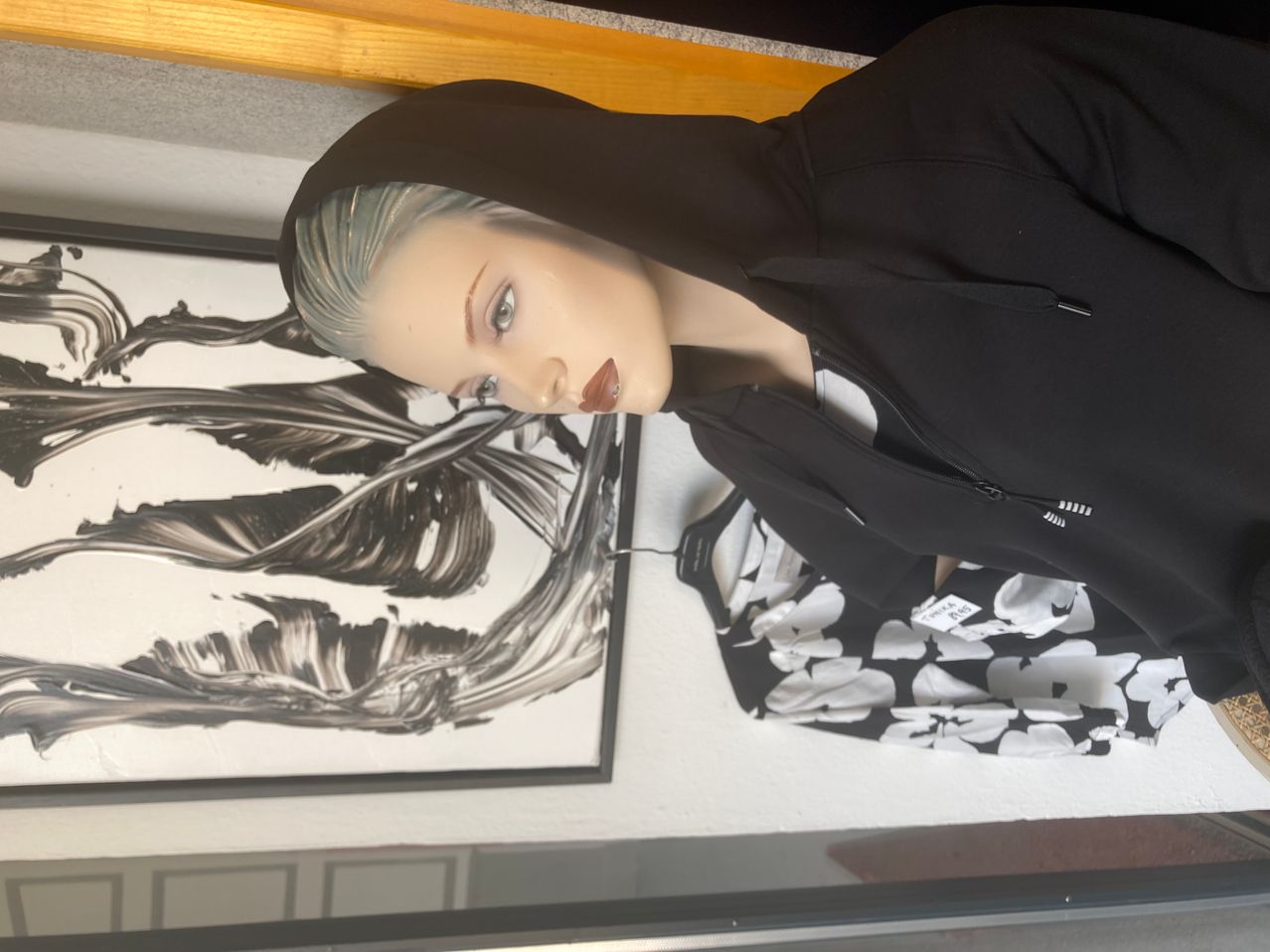 Eine Puppe in einem schwarzen Kapuzenpullover mit Kapuze steht neben einem schwarz-weißen abstrakten Gemälde. Die Puppe hat kurzes grünes Haar und Make-up.