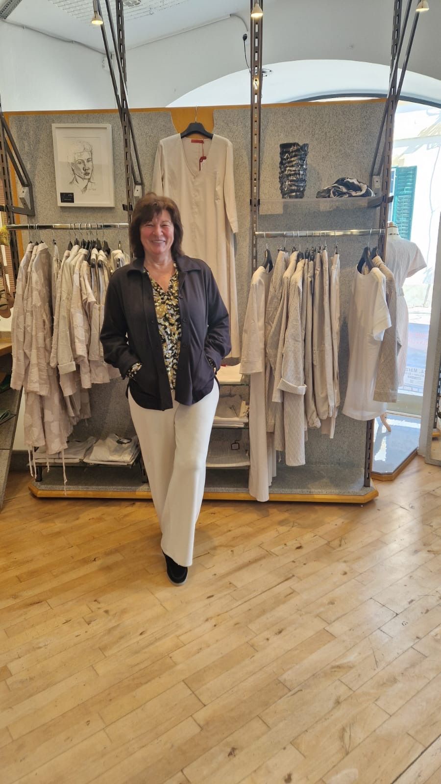 Eine Frau steht lächelnd vor einem Bekleidungsgeschäft. Sie trägt eine schwarze Jacke und weiße Hosen. Dahinter zeigt ein Kleiderständer weiße und beige Kleidungsstücke.