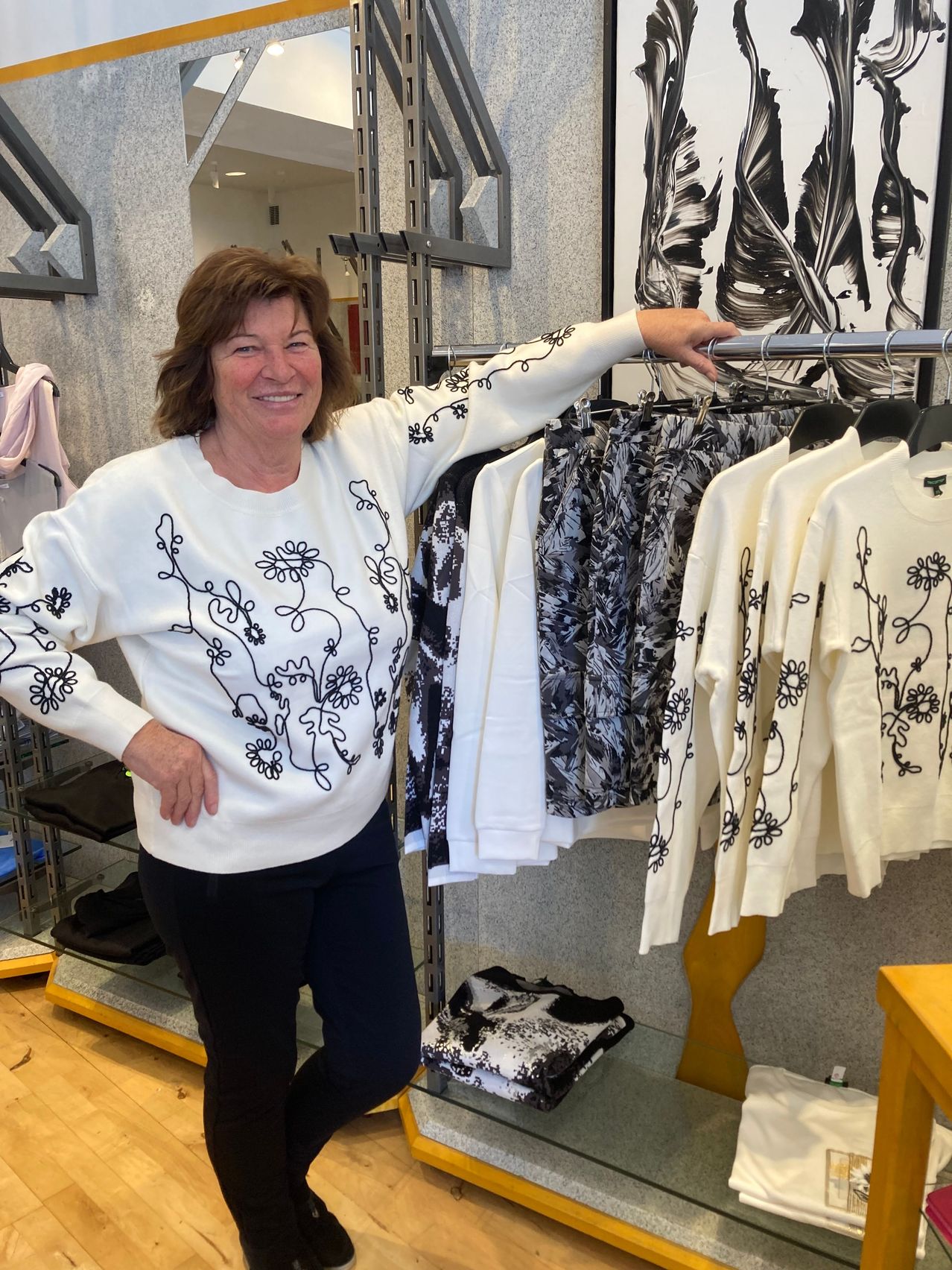 Eine Frau in einem weißen Blumenpullover steht vor einem Kleiderständer, lächelt und hält sich mit einer Hand am Ständer fest.