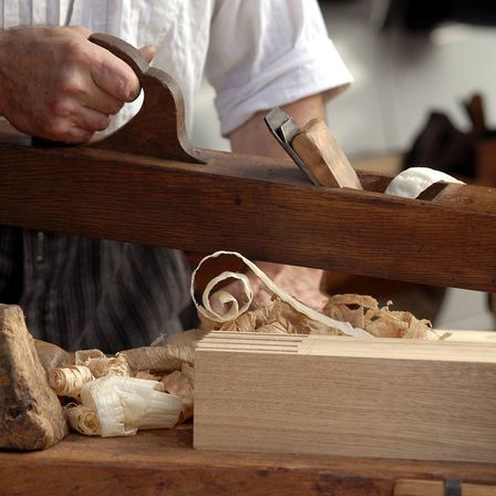Eine Person benutzt eine große hölzerne Hobelmaschine, um Holzspäne von einem Holzblock auf einem Tisch zu schälen.