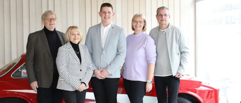 Fünf Menschen stehen lächelnd vor einem roten Auto. Sie sind formell gekleidet, der Mann in der Mitte trägt einen hellblauen Anzug. Die Frau links trägt eine gestreifte Jacke und die Frau in der Mitte einen lila Pullover. Das Auto hat ein Logo auf der Motorhaube.