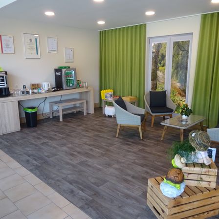 Ein Raum mit Holzboden und weißen Wänden, grünen Vorhängen, einem Tisch mit einer Kaffeemaschine, einer Bank und Stühlen. Es gibt Topfpflanzen, Skulpturen und einen Mülleimer.