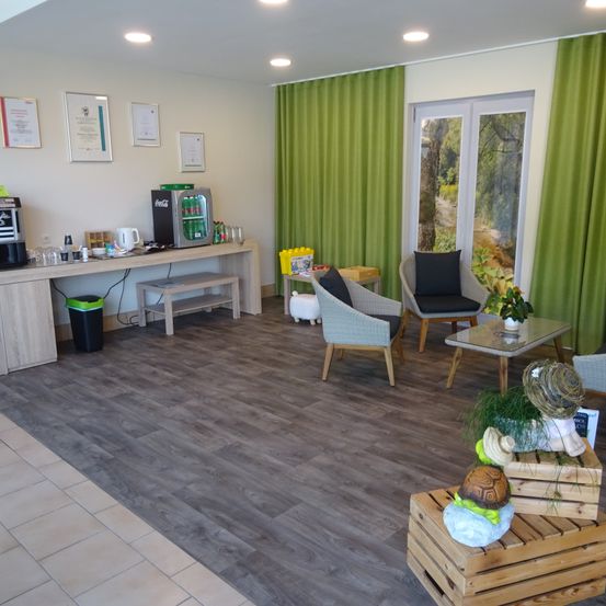 Ein Raum mit Holzboden und weißen Wänden, grünen Vorhängen, einem Tisch mit einer Kaffeemaschine, einer Bank und Stühlen. Es gibt Topfpflanzen, Skulpturen und einen Mülleimer.