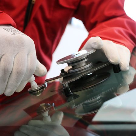 Eine Person in einem roten Jacke und weißen Handschuhen arbeitet mit einem Werkzeug an einer Glasscheibe.