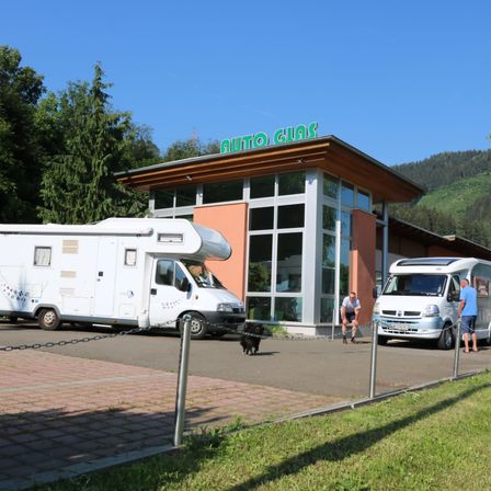 Zwei weiße Wohnmobile parken vor einem Gebäude mit einem grünen Schild, das 'Auto Gias' steht. Ein Mann und ein Hund gehen auf dem Bürgersteig.