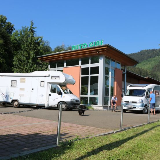 Zwei weiße Wohnmobile parken vor einem Gebäude mit einem grünen Schild, das 'Auto Gias' steht. Ein Mann und ein Hund gehen auf dem Bürgersteig.