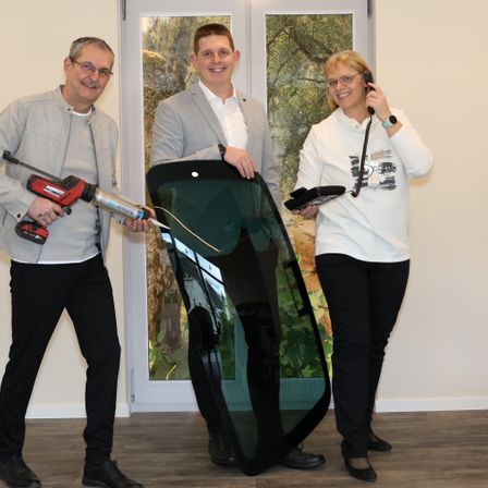 Drei Personen stehen in einem Büro, halten ein Fenster und eine Bohrmaschine. Zwei tragen Brillen. Die Frau rechts telefoniert.