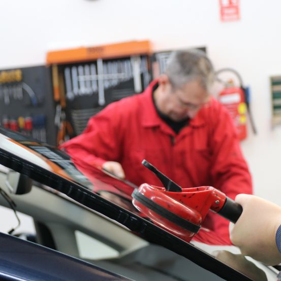 Ein Mann in einem roten Jackett arbeitet mit einem Werkzeug an der Windschutzscheibe eines Autos. Ein Feuerlöscher ist an der Wand hinter ihm montiert.
