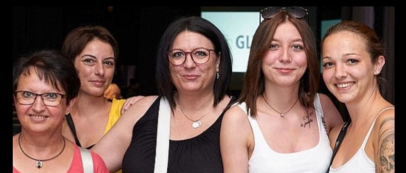 Drei Frauen stehen lächelnd zusammen. Die mittlere Frau trägt eine Brille und eine Halskette. Die Frau rechts von ihr trägt ein weißes Tanktop und hat eine Sonnenbrille auf dem Kopf. Die Frau links von ihr trägt ein gelbes Oberteil.