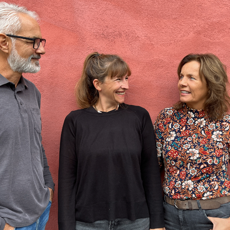 Drei Personen stehen gemeinsam vor einer roten Wand, alle unterschiedlich gekleidet. Der Mann trägt ein graues Poloshirt und hat eine Brille und einen weißen Bart. Die Frau rechts trägt ein Blumenhemd und Jeans. Die Frau in der Mitte trägt ein schwarzes Shirt und hat einen Pferdeschwanz.