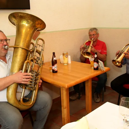 Drei Männer sitzen in einem Raum und spielen Blechblasinstrumente. Sie sitzen an einem Tisch mit Bierflaschen.