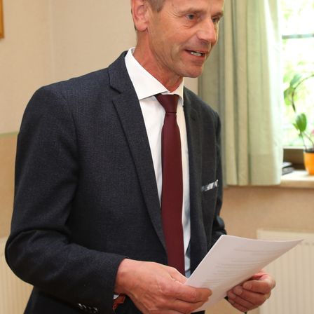 Ein Mann in einem formellen Anzug lächelt, während er ein Stück Papier in der Hand hält und vor einem Fenster mit einem Topf steht.