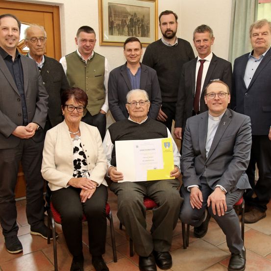 Eine Gruppe von Erwachsenen steht hinter einem sitzenden Mann, der ein Zertifikat in der Hand hält. Eine Frau sitzt neben ihm auf einem Stuhl.