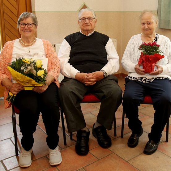 Drei ältere Erwachsene sitzen auf roten Stühlen. Die Frau links hält einen Strauß gelber Blumen, der Mann in der Mitte trägt eine Brille und eine Uhr und die Frau rechts hält einen Strauß roter Blumen.