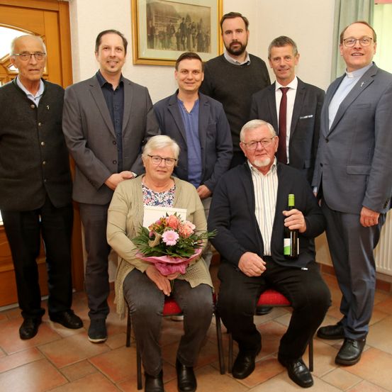 Eine Gruppe von Menschen in Anzügen posiert für ein Foto. Eine Frau in der Mitte hält einen Blumenstrauß. Ein Mann auf der rechten Seite hält eine Weinflasche.