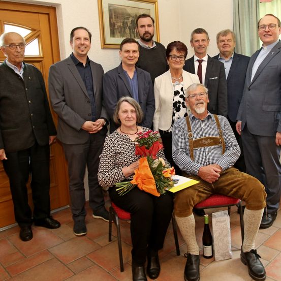 Gruppenfoto von Menschen in formeller Kleidung, die stehen und sitzen, Blumen halten und für eine Kamera lächeln.