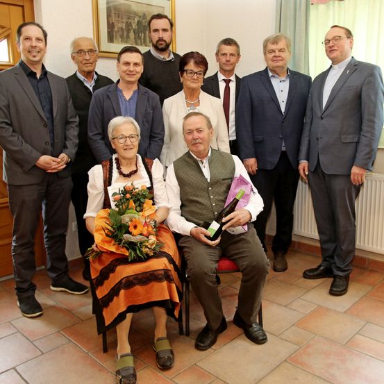 Eine Gruppe von Menschen steht in einem Raum und posiert für ein Foto. Eine ältere Frau hält einen Blumenstrauß. Ein älterer Mann sitzt auf einem Stuhl und hält eine Flasche. Ein Heizkörper steht hinter ihnen.