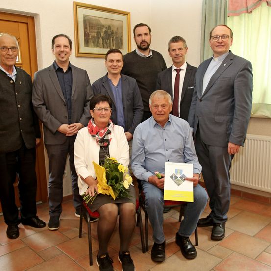 Sieben Personen stehen und sitzen in einem Raum. Ein Mann hält ein Buch und einen Stift. Die Frau sitzt auf einem Stuhl und hält einen Blumenstrauß. Dahinter ist ein Bilderrahmen an der Wand angebracht.
