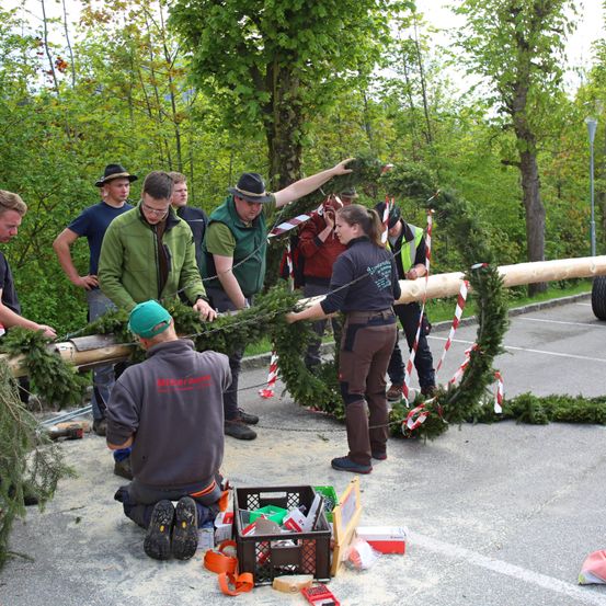 Eine Gruppe von Menschen arbeitet gemeinsam an der Herstellung eines Kranzes aus Zweigen und Grün. Sie befinden sich in einem Waldgebiet.