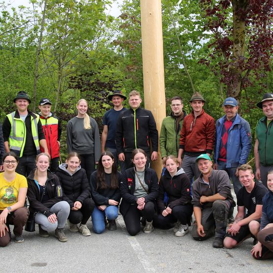 Eine Gruppe von Menschen in verschiedenen Jacken und Mützen posiert für ein Foto im Wald. Ein großer Baumstamm ist im Hintergrund zu sehen.