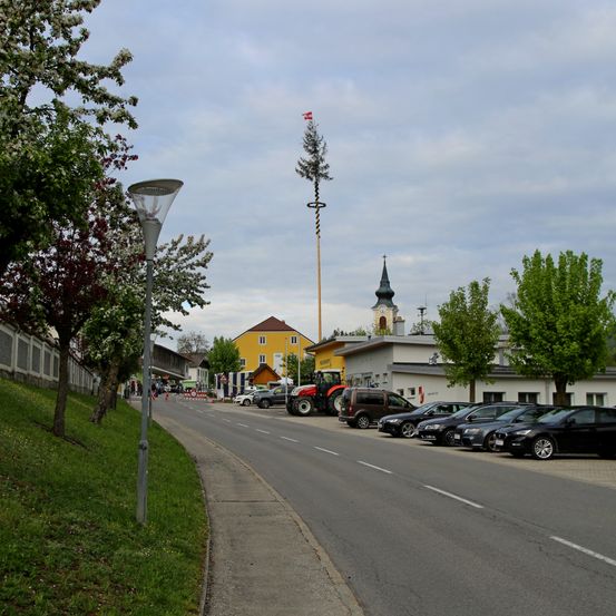 Eine Straße mit einem Bürgersteig, einer Straßenlaterne und geparkten Autos an der Seite. Es gibt eine hohe Struktur mit einer Flagge oben, und ein roter Traktor ist in der Nähe geparkt. Gebäude befinden sich auf beiden Seiten der Straße.