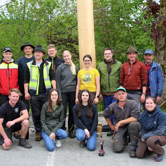Eine Gruppe junger Erwachsener posiert für ein Foto in einem Wald mit einem großen Holzpfahl hinter ihnen.