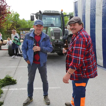 Zwei Männer stehen vor einem Traktor. Einer trägt ein kariertes Hemd und der andere ein blaues Jackett. Sie lächeln beide. Im Hintergrund sind Menschen und Bäume zu sehen.