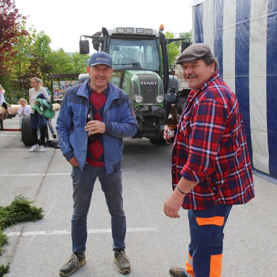 Zwei Männer stehen vor einem Traktor. Einer trägt ein kariertes Hemd und der andere ein blaues Jackett. Sie lächeln beide. Im Hintergrund sind Menschen und Bäume zu sehen.