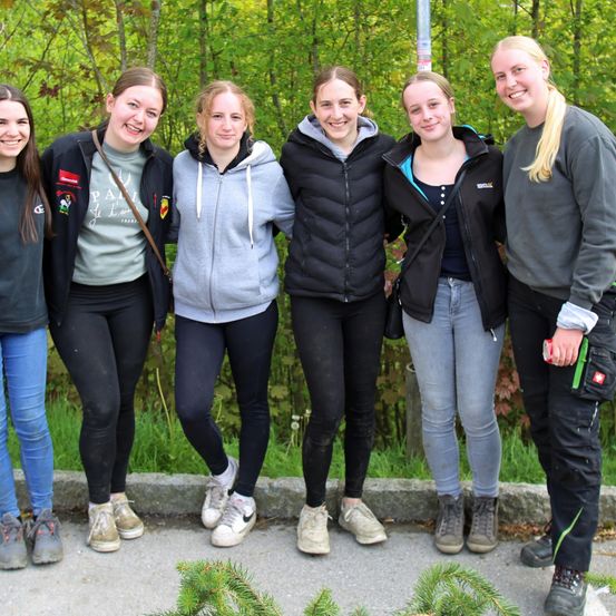 Fünf Frauen stehen vor einem Wald. Sie alle lächeln und scheinen ein Foto zu machen.