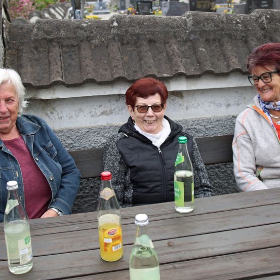 Drei Frauen sitzen auf einer Bank auf einem Friedhof. Sie lächeln und haben Flaschen mit Saft vor sich.