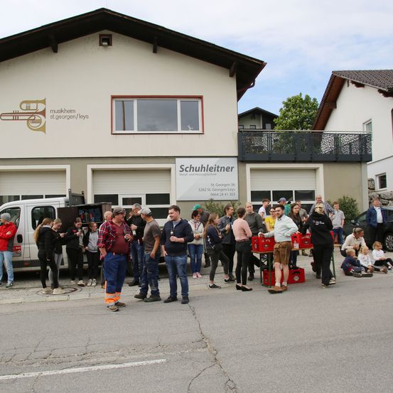 Menschen versammeln sich auf der Straße vor einem Gebäude namens Schuhleitner. Es sitzen Kinder auf dem Boden, ein weißer Lieferwagen ist geparkt und ein Banner steht auf dem Bürgersteig.