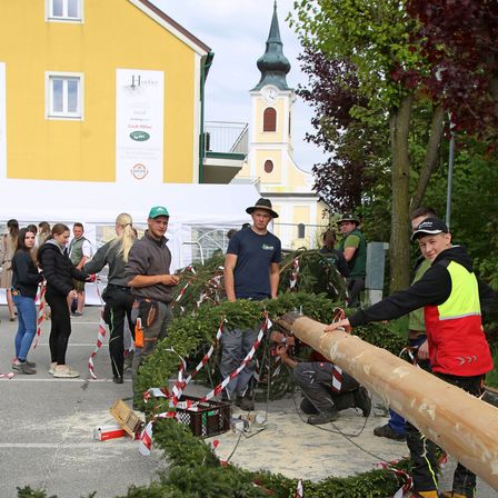 Eine Gruppe von Menschen ist im Freien. Eine Person hält einen langen Holzstamm. Sie stellen einen Kranz aus Tannenzweigen her.