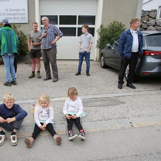 Drei Erwachsene und drei Kinder stehen auf einem Betonsteig vor einem Gebäude. Die Erwachsenen unterhalten sich miteinander, während die Kinder auf dem Bürgersteig sitzen.