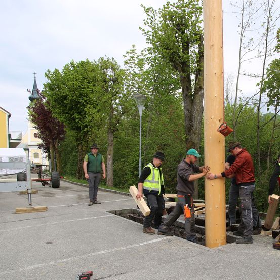 Eine Gruppe von Männern baut einen Holzpfahl auf einem Parkplatz. Ein Mann hält eine Bohrmaschine, die anderen halten Holzbretter. Dahinter steht eine Straßenlaterne und ein Gebäude.