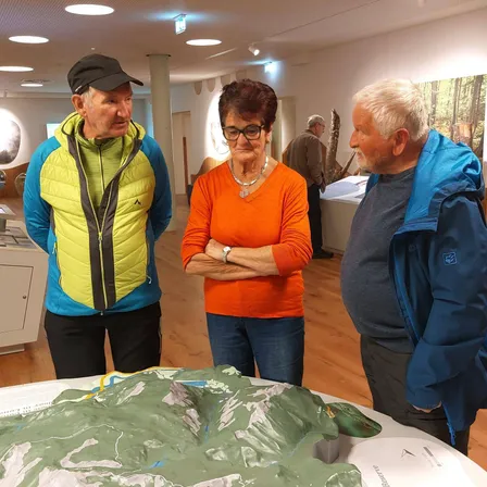 Drei Personen stehen neben einer großen topografischen Karte in einem Museum. Der Mann in der gelben und blauen Jacke schaut die Frau in Orange an. Hinter ihnen steht ein Mann in einer blauen Jacke und lauscht.