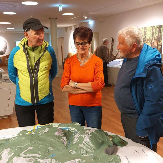 Drei Personen stehen neben einer großen topografischen Karte in einem Museum. Der Mann in der gelben und blauen Jacke schaut die Frau in Orange an. Hinter ihnen steht ein Mann in einer blauen Jacke und lauscht.