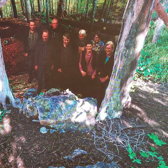 Eine Gruppe von Personen steht in einer Waldlichtung, einige von ihnen tragen Brillen. Ein großer Felsen ist im Vordergrund, und sie scheinen für ein Foto zu posieren.