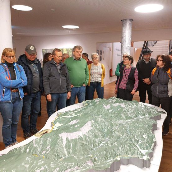 Eine Gruppe von Menschen steht um eine große, grüne Karte einer Bergkette in einer Kunstgalerie. Sie tragen alle Winterkleidung.