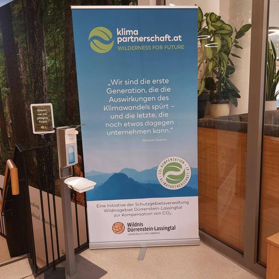 Ein blauer Banner mit Waldhintergrund sagt 'Wir sind die erste Generation, die die Auswirkungen des Klimawandels spürt - und die letzte, die noch etwas dagegen unternehmen kann.' - Barack Obama. Es ist eine Initiative zur CO2-Kompensation.