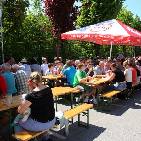 Eine Menschenmenge sitzt unter einem roten Regenschirm an Tischen und genießt Getränke und Essen. Eine Frau hält ein Baby auf dem Schoß. Bäume und eine Straßenlaterne sind im Hintergrund.
