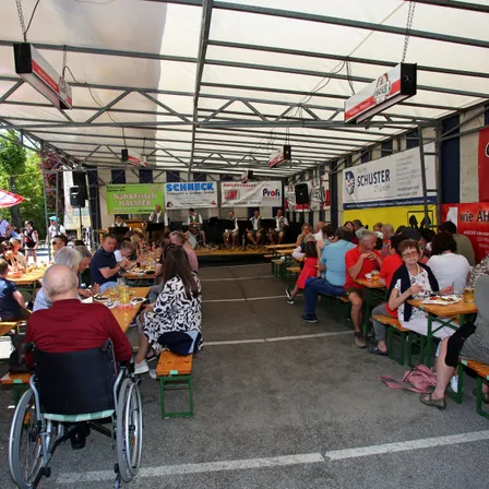 Eine Gruppe von Menschen speist unter einem zeltgedeckten Pavillon, mit Musikern auf der Bühne im Hintergrund. Die Tische sind in Reihen angeordnet, und es hängen Banner von der Decke.