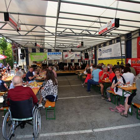 Eine Gruppe von Menschen speist unter einem zeltgedeckten Pavillon, mit Musikern auf der Bühne im Hintergrund. Die Tische sind in Reihen angeordnet, und es hängen Banner von der Decke.
