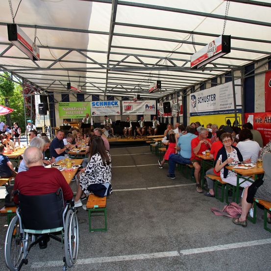 Eine Gruppe von Menschen speist unter einem zeltgedeckten Pavillon, mit Musikern auf der Bühne im Hintergrund. Die Tische sind in Reihen angeordnet, und es hängen Banner von der Decke.