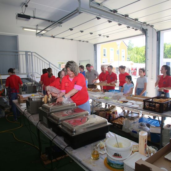 Eine Gruppe von Menschen in roten T-Shirts bereitet Essen in einer Garage vor. Sie stehen um einen langen Tisch mit Tabletts mit Essen, Schüsseln und Behältern herum. Es gibt andere Leute, die herumstehen und zuschauen.